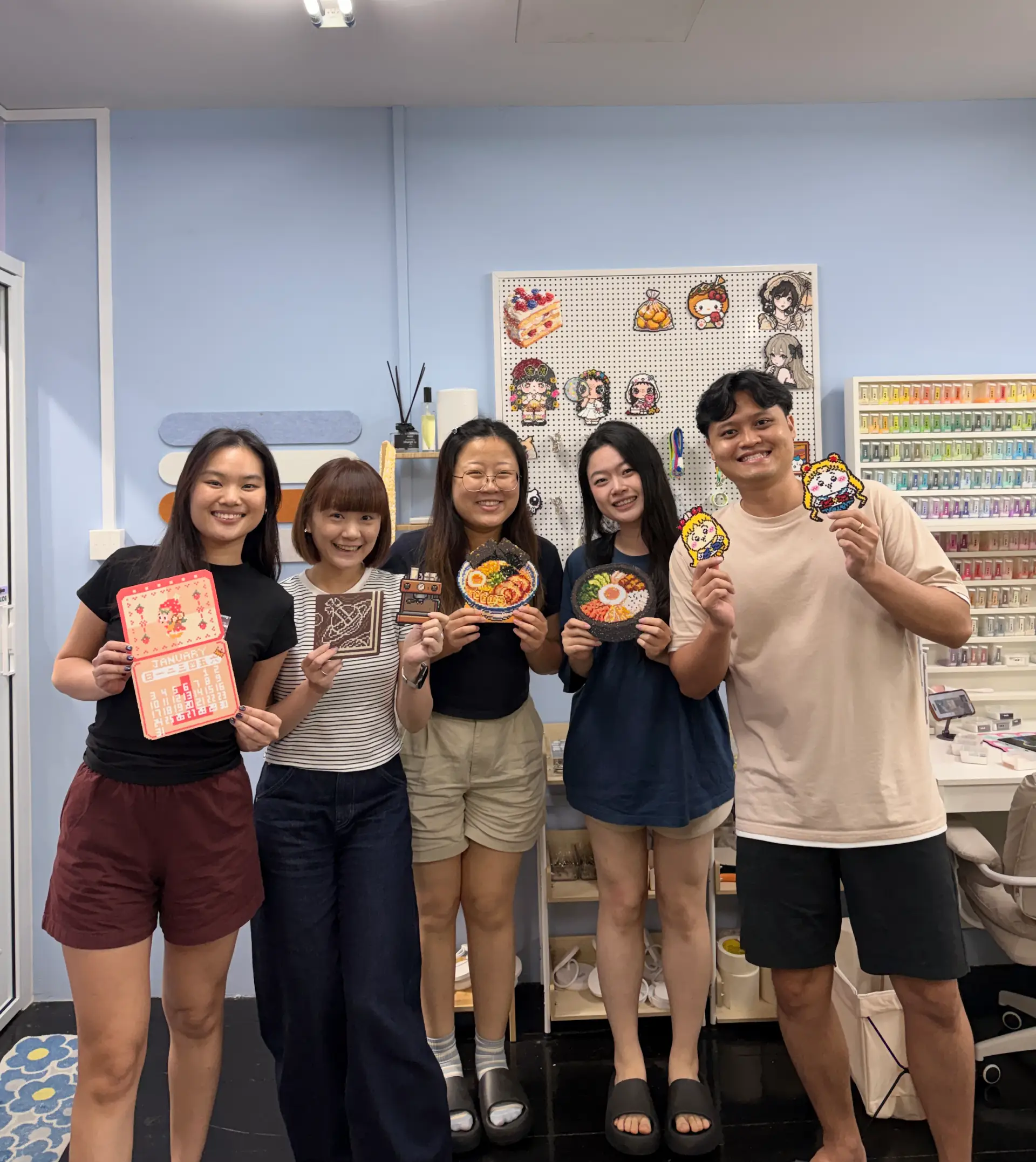 Five friends in the studio holding up the bead pieces they made, in front of the pegboard wall of finished designs